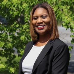 Photo of Angela Johnson standing outside in front of a tree. She is wearing a white shirt under a black striped blazer