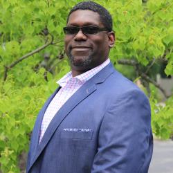 Photo of Anwar Young outside in front of a tree. He is wearing a pale blue suit jacket with a light pink button down and black sunglasses. Anwar has short dark hair. 