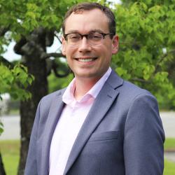 Photo of Ben standing outside in front of a tree. Ben is wearing glasses, has short brown hair, grey jacket and pink button up shirt