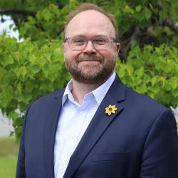 photo of DJ Hampton outside in front of a tree. DJ is wearing glasses, dark blue suit jacket, blue plaid button up, yellow sunflower on the suit jacket lapel