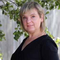 Ilze has shoulder length blonde hair, and is wearing a black top. She is standing outside in front of a tree and fence