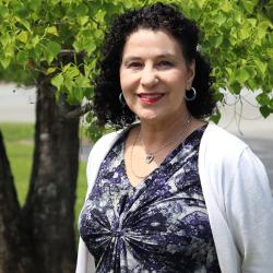 Photo of Joyce standing in front of tree, wearing a white cardigan and patterned top. Joyce has dark, curly hair