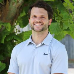 Headshot photo of Justin McCloskey, wearingshort sleeve blue polo shirt, smiling, standing in front of tree outside