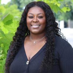 Photo of Kayela Rose outside in front of a tree. She is wearing a black long sleeve blouse. Kayela has curly long dark hair. 