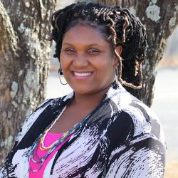 Photo of La Toya Jefferson outside in front of a tree. She is wearing a hot pink shirt with a marble pattern coat and colorful beaded necklaces. LaToya has her hair up in braids and is smiling. 