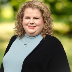 Headshot photo of Andrea Boccucci in front of a tree. She is wearing a light blue turtle neck with a black cardigan. Andrea has short curly blonde hair. 