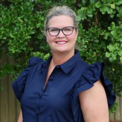 Photo of Lee Anne Stone, she is standing outside in front of a tree, smiling, wearing glasses, hair pulled back and a blue dress with puffy sleeves