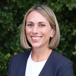 Photo of Devon Wade outside in front of a tree with green leaves. She is smiling, has shoulder length hair, wearing a white top and a navy blue blazer