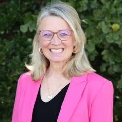 Julie Frye is standing outside in front of a tree with green leaves. She is smiling, wearing glasses, black top and a pink blazer.