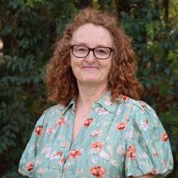 Kimberly wears glasses and a light blue floral dress. She has shoulder length red curly hair.