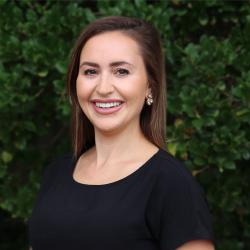 Sarah Makuta is posing outside in front of a tree with green leaves wearing a black short sleeve top.