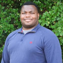 Shaq Jordan is standing outside in front of a green tree. He is wearing a longsleeve blue polo shirt and is smiling at the camera.