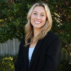 Emily is standing outside, smiling at the camera. She has long blonde hair and wears a dark blazer and white top.