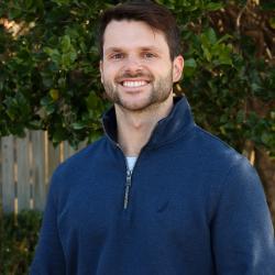 Headshot photo of Justin McCloskey, wearing long sleeve dark three-quarter zip, smiling, standing in front of tree outside