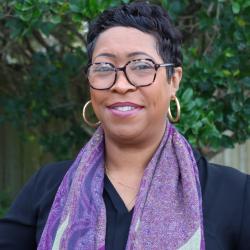 Photo of Khalilah Sumners, she is standing outside in front of a tree. She wears glasses, a black top and purple scarve. She has short hair and is smiling, looking at the camera.