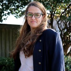 Kristie is standing outside, she has long blonde hair and wears glasses. She has on a white top with a blue blazer, and is smiling at the camera