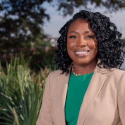 Photo of Natasha Chatman. Natasha is African American and has shoulder-length black curly hair, she is smiling and looking at the camera.  Natasha is wearing a bright green top and beige blazer, she is standing outside.